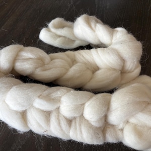 May include: Two braids of soft, white wool roving, ideal for fiber art projects. The wool is thick and fluffy, with a slightly textured appearance. The braids are arranged on a dark brown surface.
