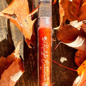 May include: A clear lip gloss tube filled with orange-red liquid and glitter, with the words "Roselight Enchantments" in cursive script. The tube is surrounded by fallen orange autumn leaves.