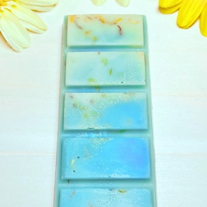 Puede incluir: Una barra de cera para fundir azul y blanca con cinco cuadrados individuales. La barra de cera para fundir est&aacute; sobre una superficie blanca con flores amarillas.