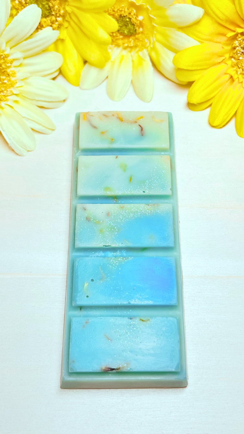 Puede incluir: Una barra de cera para fundir azul y blanca con cinco cuadrados individuales. La barra de cera para fundir est&aacute; sobre una superficie blanca con flores amarillas.