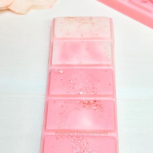 May include: A pink wax melt bar with four sections, each a different shade of pink. The top section is a light pink with gold flecks, and the bottom section has the text "Roselight Enchantments".
