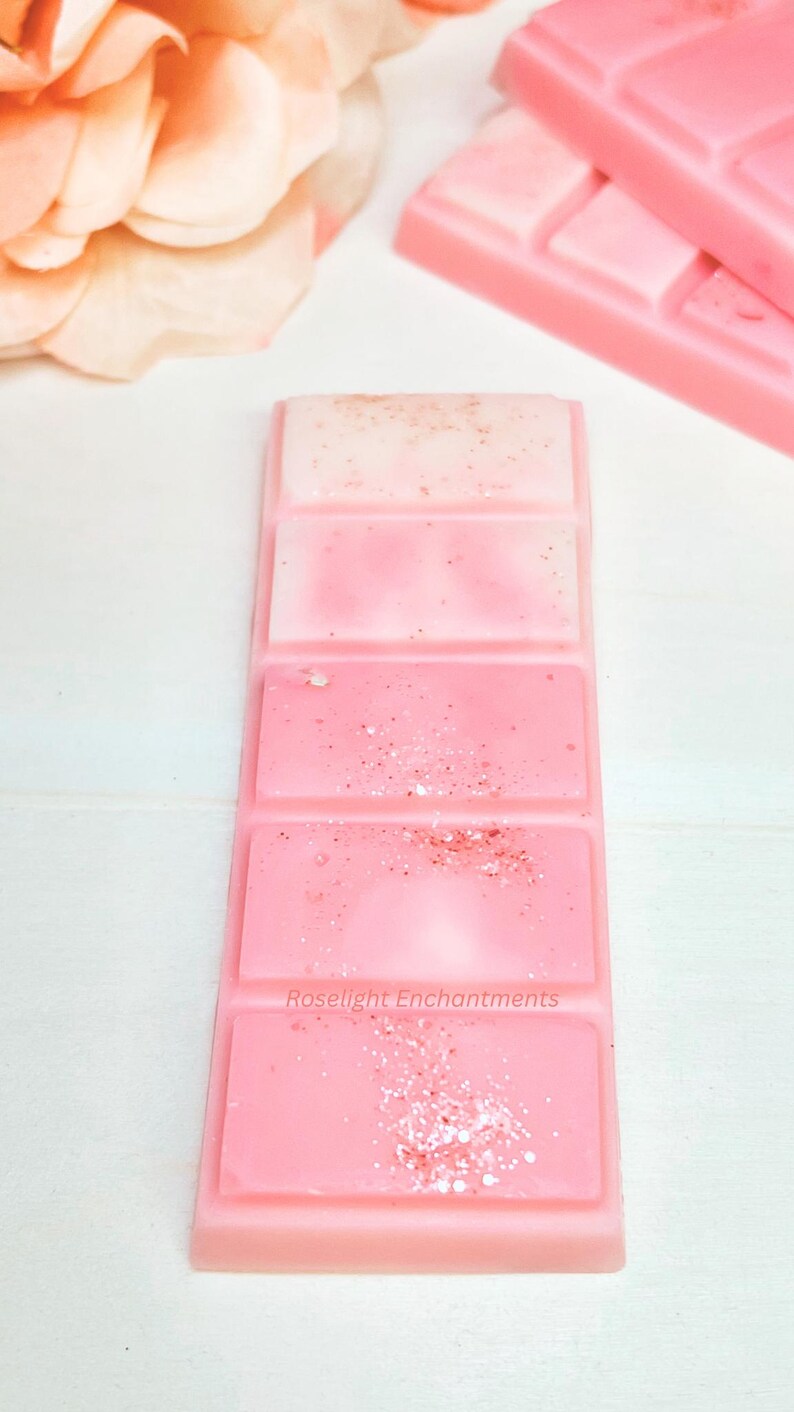 May include: A pink wax melt bar with four sections, each a different shade of pink. The top section is a light pink with gold flecks, and the bottom section has the text "Roselight Enchantments".