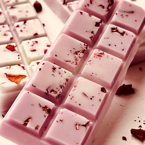 Puede incluir: Una barra de cera fundida de color lavanda con un patr&oacute;n de cuadr&iacute;cula y p&eacute;talos de rosa secos. La barra est&aacute; dividida en cuadrados y tiene un fondo blanco.