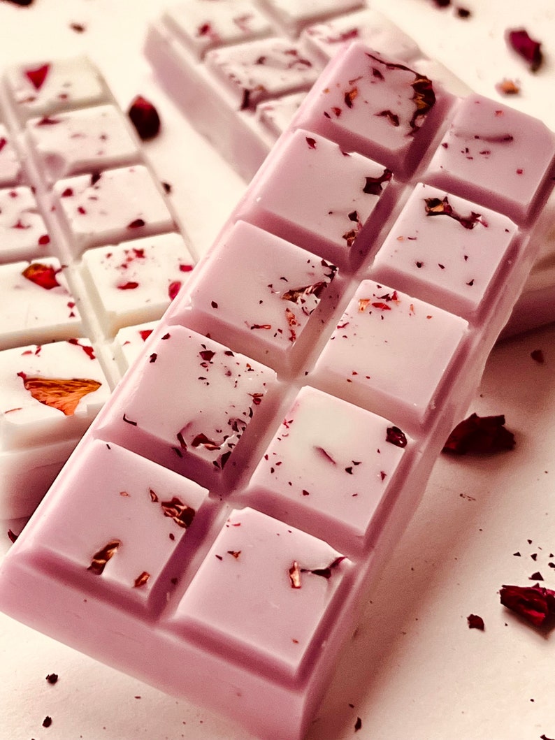 Puede incluir: Una barra de cera fundida de color lavanda con un patr&oacute;n de cuadr&iacute;cula y p&eacute;talos de rosa secos. La barra est&aacute; dividida en cuadrados y tiene un fondo blanco.