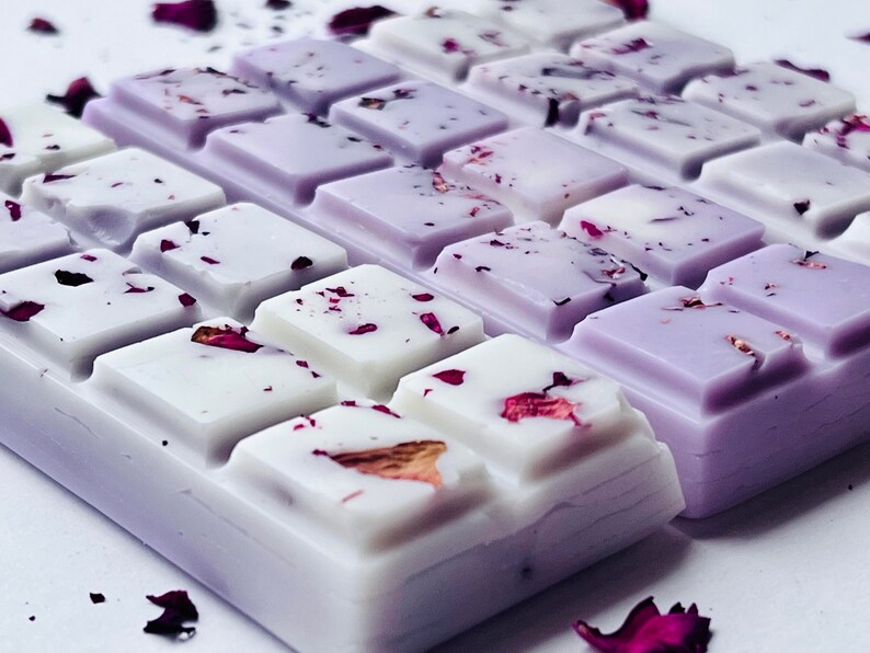 May include: Close-up of wax melts in a square shape, divided into rectangular sections. The wax is white and lavender, with dried rose petals sprinkled on top. The arrangement suggests a spa or aromatherapy product.