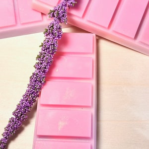 May include: Pink wax melt bars with four rectangular sections, arranged with sprigs of purple, beaded floral stems. The wax melts are a soft pink color, and the floral stems are a deep purple with green accents. The background is a light wood grain.