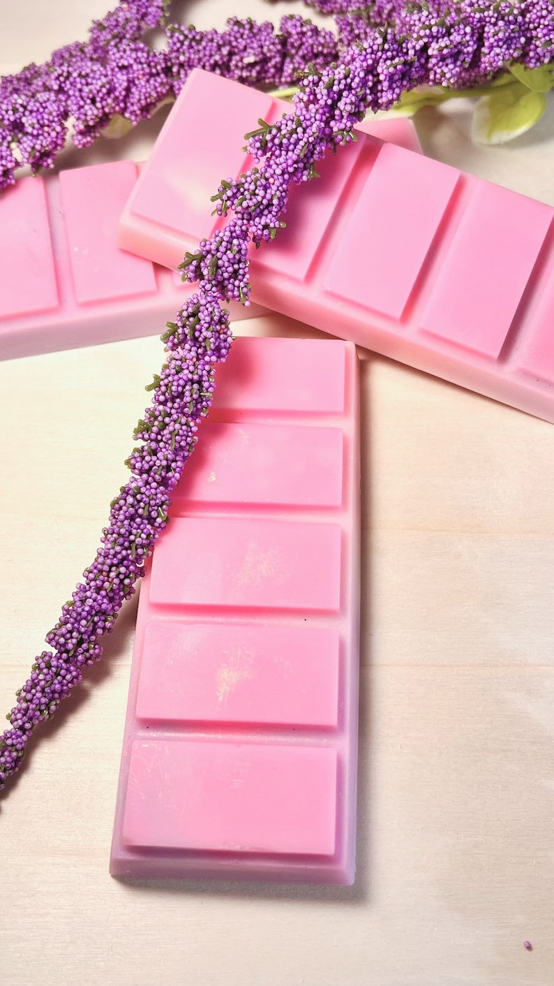 May include: Pink wax melt bars with four rectangular sections, arranged with sprigs of purple, beaded floral stems. The wax melts are a soft pink color, and the floral stems are a deep purple with green accents. The background is a light wood grain.