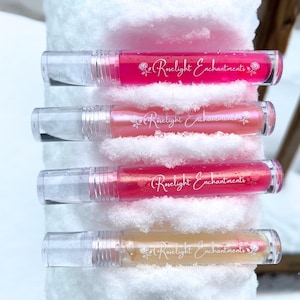 Könnte beinhalten: Vier Lipgloss-Tuben in Rosa- und Pfirsichtönen mit transparenten Kappen liegen auf Schnee. Jede Tube ist mit "Roselight Enchantments" in Schreibschrift beschriftet. Der Hintergrund zeigt eine verschneite Szene im Freien.