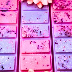 May include: Rows of pink and purple wax melts with embedded dried botanicals and glitter. The wax melts are rectangular and arranged in a grid pattern. The text "Roselight Enchantments" is visible on one of the wax melts. A pink flower is at the top.