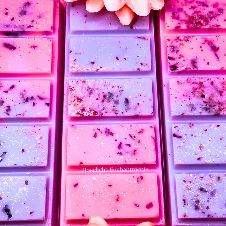 May include: Rows of pink and purple wax melts with embedded dried botanicals and glitter. The wax melts are rectangular and arranged in a grid pattern. The text "Roselight Enchantments" is visible on one of the wax melts. A pink flower is at the top.