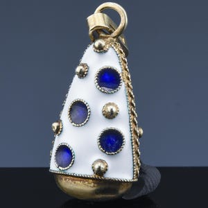 May include: A gold-toned pendant with a white enamel surface, adorned with blue circular accents and gold embellishments. The pendant has a bell-like shape with a loop at the top for hanging. The edges are trimmed with a gold chain.
