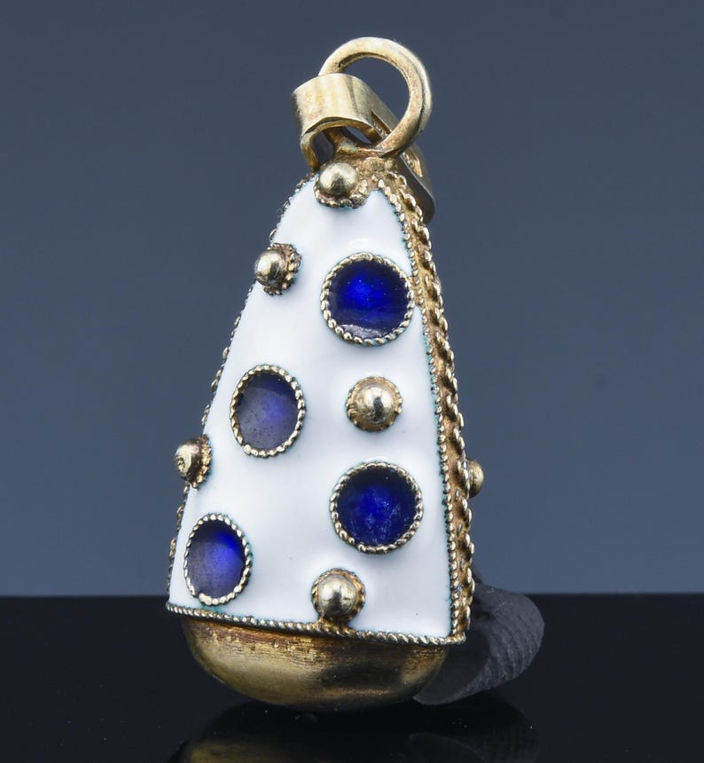 May include: A gold-toned pendant with a white enamel surface, adorned with blue circular accents and gold embellishments. The pendant has a bell-like shape with a loop at the top for hanging. The edges are trimmed with a gold chain.