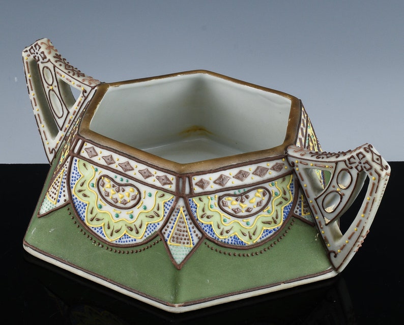 May include: A hexagonal ceramic dish with two ornate handles. The dish is decorated with a green, brown, and yellow geometric pattern.