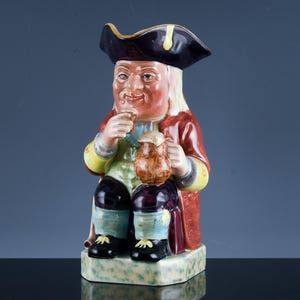 May include: A ceramic Toby jug figurine of a seated man in a tricorn hat and red coat, holding a mug and a small object. The jug is hand-painted with a detailed design and rests on a light green base.