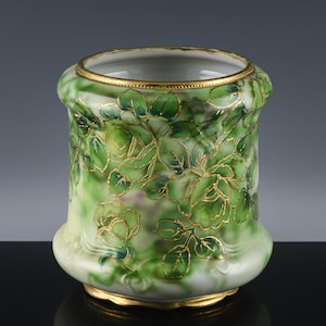 May include: A cylindrical ceramic vase with a green and white floral design. The vase features gold accents on the rim, base, and outlining the floral pattern. The flowers are a mix of green shades, creating a detailed and elegant look.