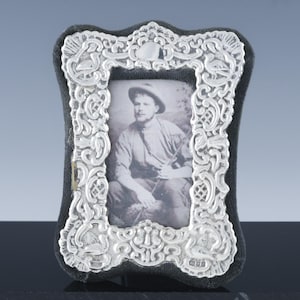 May include: A silver-colored, ornate picture frame with a black velvet backing. The frame has a detailed floral and scroll design. The photograph inside is a black and white portrait of a man wearing a hat. The frame is approximately 5 inches tall.