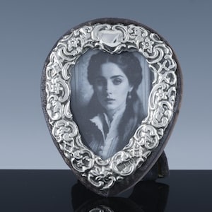 May include: A heart-shaped silver picture frame with an ornate floral design. The frame holds a black and white portrait of a woman. The frame has a dark backing and stands upright on a black surface.
