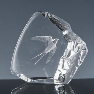 May include: A clear glass sculpture featuring a detailed carving of a bird in flight. The sculpture has a rough, textured edge on one side, and a smooth, polished surface. The bird's wings are outstretched, and the sculpture is set against a dark background.