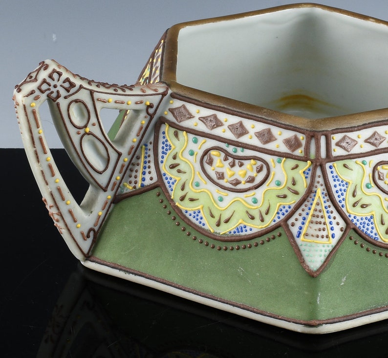 May include: A hexagonal ceramic sugar bowl with a green and brown geometric design. The bowl has a handle and a white interior.