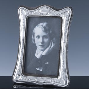 May include: A vintage silver-toned picture frame with a portrait of a person. The frame has a curved, ornate design with decorative detailing. The portrait is in black and white.