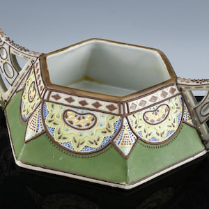 May include: A green and white porcelain sugar bowl with ornate gold and brown details. The bowl has two handles and a hexagonal shape.