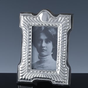 May include: A silver-colored picture frame with an ornate, detailed design. The frame holds a black and white portrait of a woman. The frame has a rectangular shape with a decorative top and bottom.