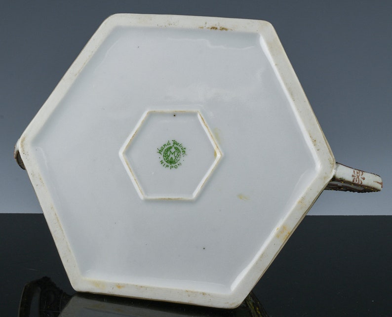 May include: The underside of a white hexagonal teapot with a green and white "Hand Painted Nippon" mark.