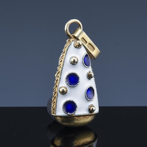 May include: A gold pendant with a white enamel surface, adorned with blue circular accents. The pendant has a gold chain border and a gold bail for attaching to a necklace. The bottom of the pendant is also gold.