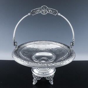 May include: A silver-colored, ornate serving basket with a handle. The basket has a round bowl shape with a decorative floral pattern around the rim. It stands on a pedestal base with three ornate feet. The handle is arched and connects to the bowl.