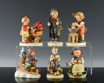 May include: A collection of six hand-painted ceramic figurines depicting children in various activities, including a child with a baby carriage, a chimney sweep, a child bathing a doll, a child on a sled, a child with geese, and a child with flowers.