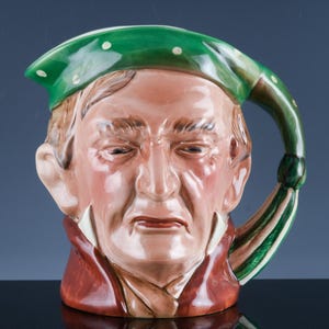 May include: A ceramic pitcher shaped like a man's head, featuring a green hat with white dots and a handle with green and white stripes. The face is detailed with realistic features and painted in shades of brown, red, and white.