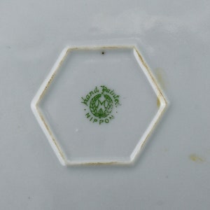 May include: Close-up of a hexagonal mark on a white ceramic plate. The mark is green and reads "Hand Painted" in a circle around a stylized "M" with a wreath. Below the circle is the word "NIPPON".