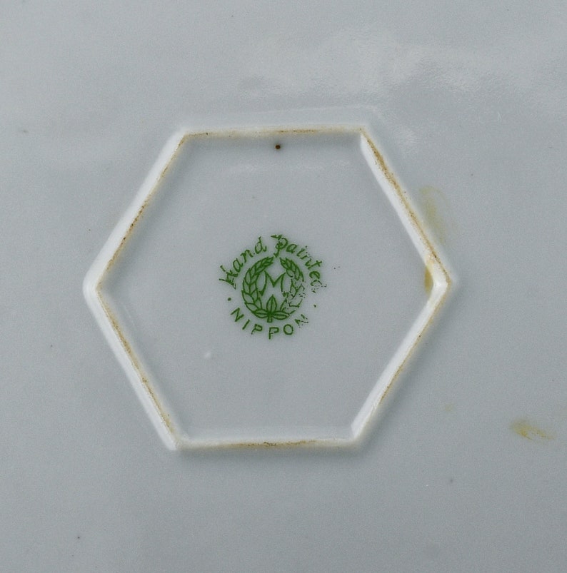 May include: Close-up of a hexagonal mark on a white ceramic plate. The mark is green and reads "Hand Painted" in a circle around a stylized "M" with a wreath. Below the circle is the word "NIPPON".