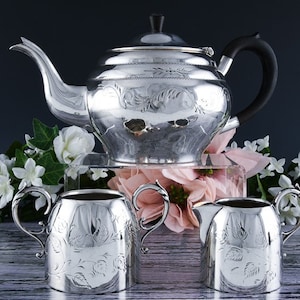 May include: A silver tea set with an ornate design. The set includes a teapot, creamer, and sugar bowl. The teapot has a black handle.