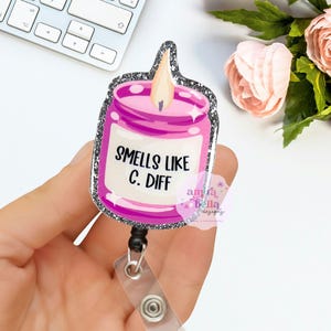 Puede incluir: Un portatarjetas en forma de vela rosa con un borde de purpurina plateada. La tarjeta de identificación dice "Smells Like C. Diff" en texto negro. La tarjeta de identificación se sujeta con un clip de plástico transparente y un cordón retráctil negro.