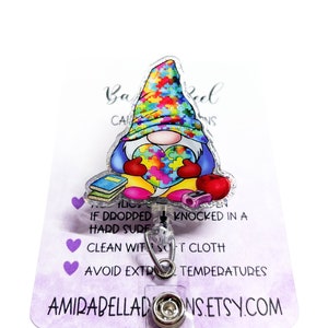 May include: A colorful puzzle piece heart gnome badge reel with a purple background. The text on the card reads "Babeel Creations, If dropped hard surface, Clean with soft cloth, Avoid extreme temperatures, AmirabellaDons.etsy.com".