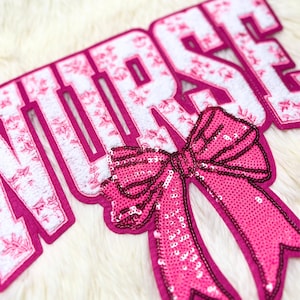 May include: A pink and white felt applique spelling "NURSE". The letters are outlined in pink and filled with a floral design. A sequined pink bow is attached below the letters, all on a fluffy white background.