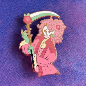 May include: A pink enamel pin featuring a stylized Grim Reaper with pink curly hair, a skull face, and a green-tinted hand. The Reaper holds a scythe with a white blade and green trim. The pin has gold accents and a rose design.