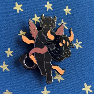 Puede incluir: Un pin de esmalte negro con una figura de querubín con cuernos y alas, abrazando a un toro. El pin tiene detalles en naranja y rosa, sobre un fondo azul con estrellas doradas. Un accesorio único.