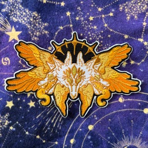May include: Embroidered patch featuring a stylized wolf with golden wings and a sunburst design. The patch has a white, gold, and black color scheme with intricate details. The patch is set against a dark blue fabric with a celestial pattern.