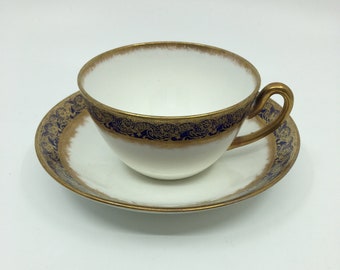 Può includere: Una tazzina da tè e un piattino bianchi con un bordo dorato e blu. La tazza e il piattino hanno un delicato motivo floreale.
