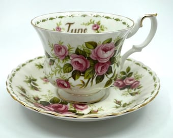 May include: A white teacup and saucer set featuring a floral rose design in pink and green. The teacup has a gold-trimmed handle and the word "June" printed on the rim. The saucer has a scalloped edge with gold trim.