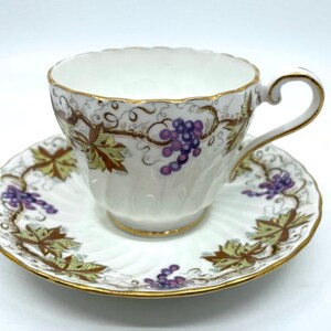 May include: A white teacup and saucer with a grapevine design. The cup and saucer have a gold trim and are decorated with purple grapes, green leaves, and brown vines.