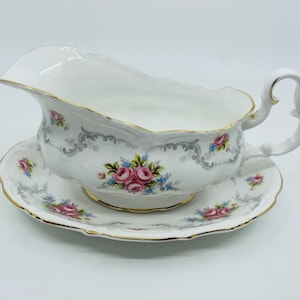 Könnte beinhalten: Eine weiße Porzellan-Soßenkännchen mit goldener Verzierung und einem Blumenmuster. Die Kanne steht auf einer passenden Untertasse.