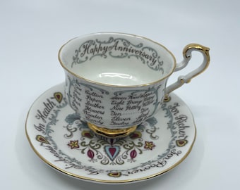 May include: A white teacup and saucer set with gold trim. The teacup has a gold handle and the saucer has a gold rim. The teacup and saucer are decorated with a floral pattern and the words "Happy Anniversary" and "In Health, For Richer, For Poorer".