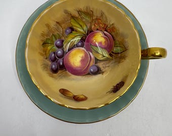 May include: A vintage teacup and saucer set. The teacup and saucer are light green with gold trim. The teacup interior features a painting of peaches and grapes. The handle of the teacup is gold.