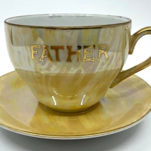 May include: A yellow teacup and saucer set with a gold iridescent glaze. The teacup has the word "FATHER" written in gold lettering.