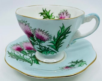 May include: A light blue teacup and saucer set with a floral design featuring pink and green thistles. The set has a gold trim around the edges.