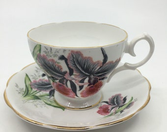 Puede incluir: Un juego de taza y platillo blanco con un diseño floral con orquídeas rosas y negras. El juego tiene un borde dorado.
