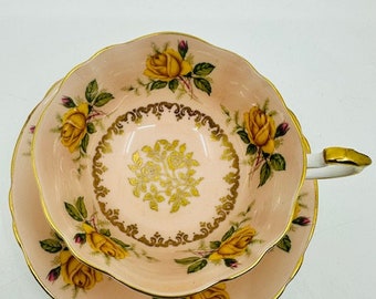 May include: A pink teacup and saucer set with a gold floral design and yellow roses. The set is made of fine china and has a delicate, feminine feel.